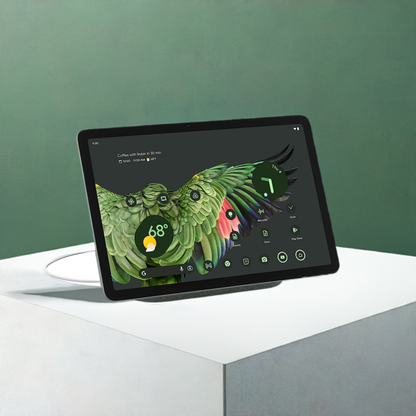 A Kosher Google Pixel Tablet displaying a vibrant wallpaper of a green parrot sits on a white stand against a green background. The screen shows a weather widget and various app icons.